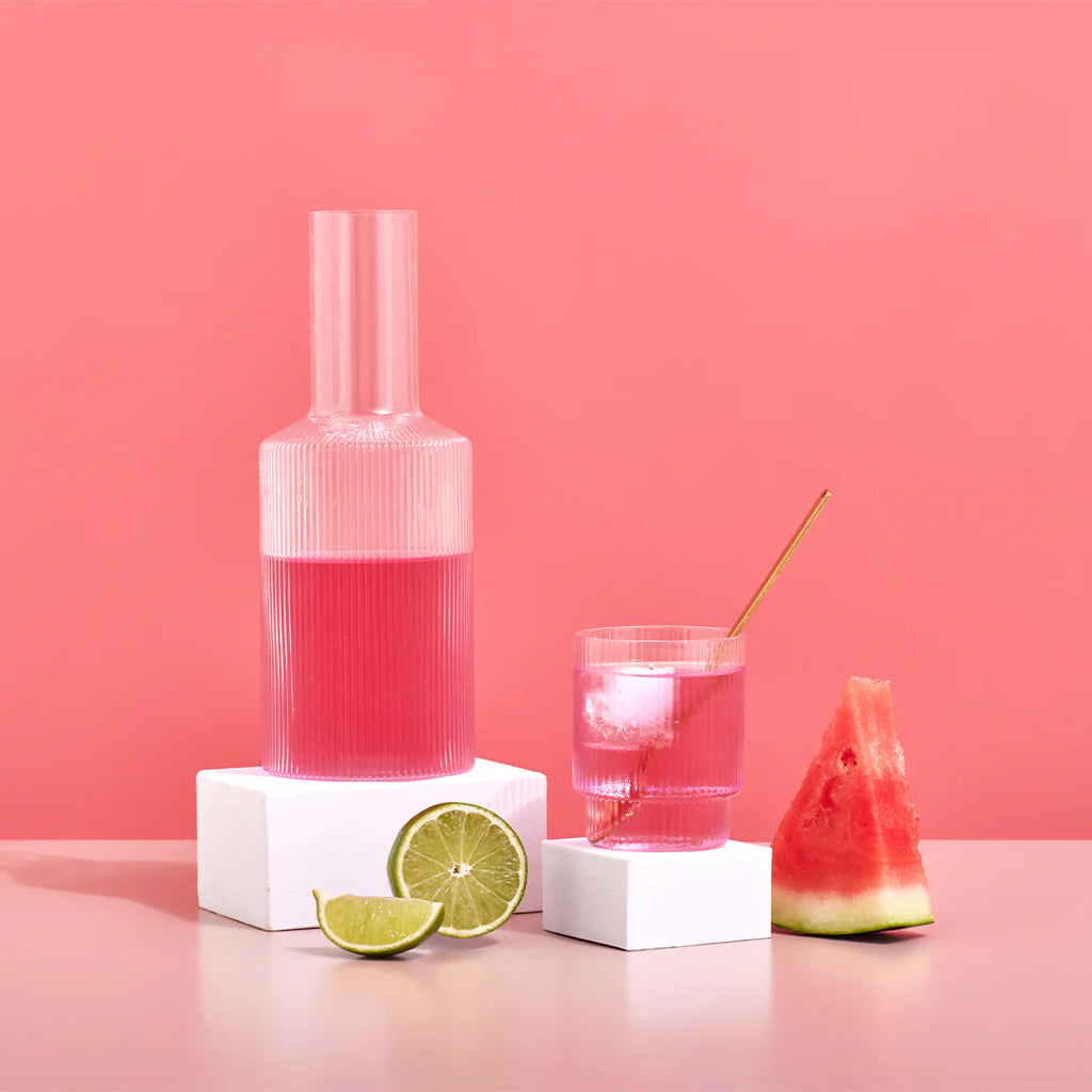 Carafe and glass filled with Hydramama Watermelon & Lime drink, styled with lime and watermelon slices on a pink backdrop.