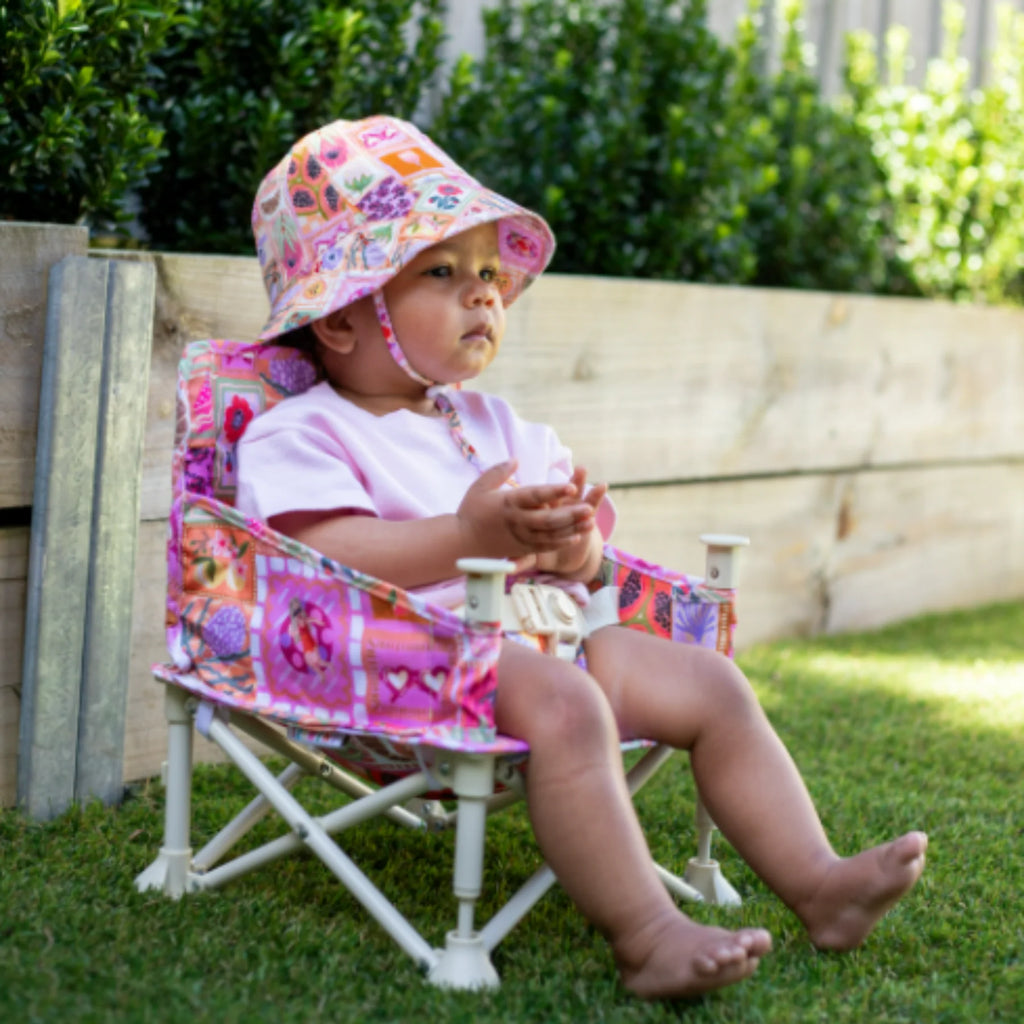 A baby wearing a matching Amy-patterned hat sits in the Izimini Baby Chair outdoors on grass.