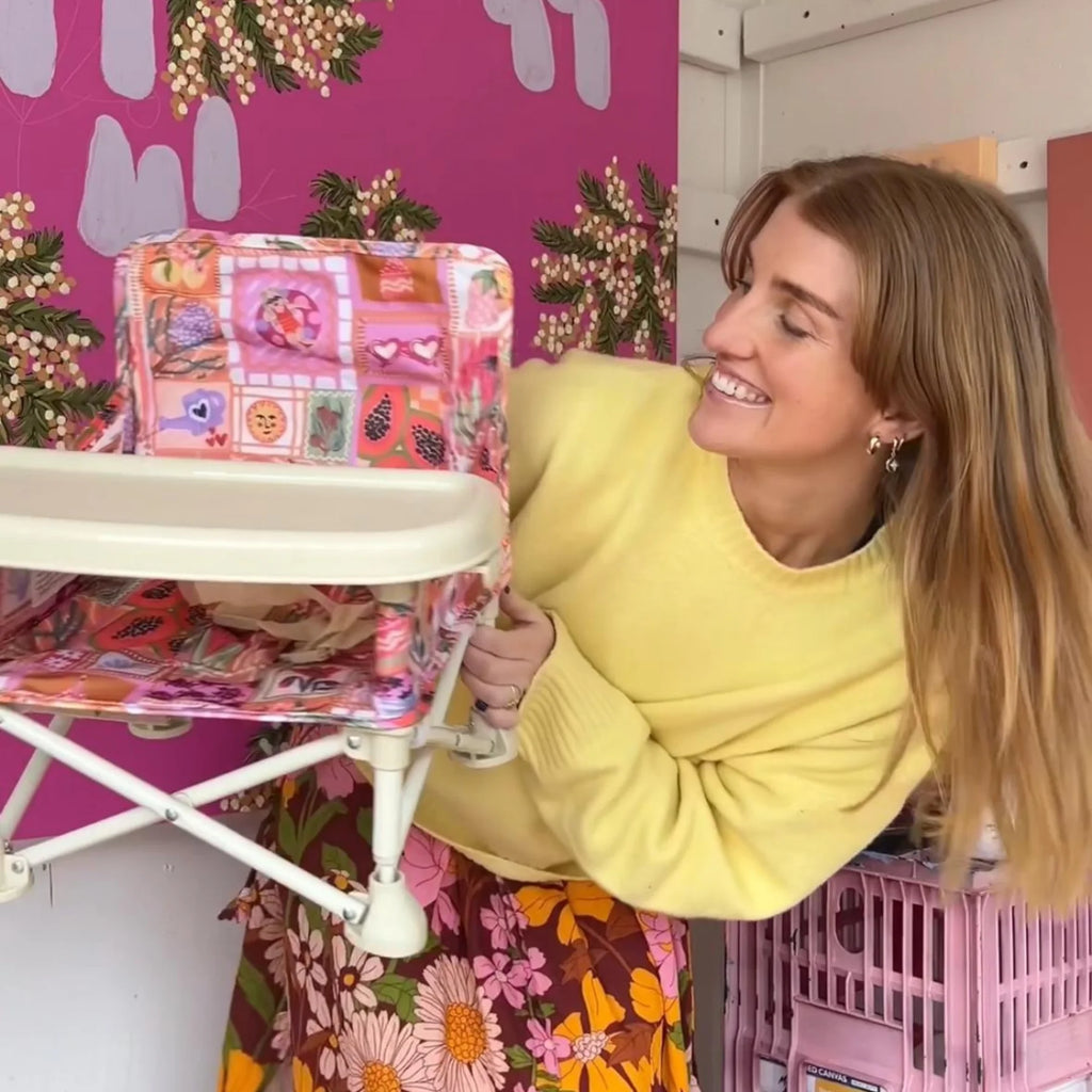 A woman in a yellow sweater and floral skirt smiles while holding the Izimini Baby Chair in the Amy print indoors.