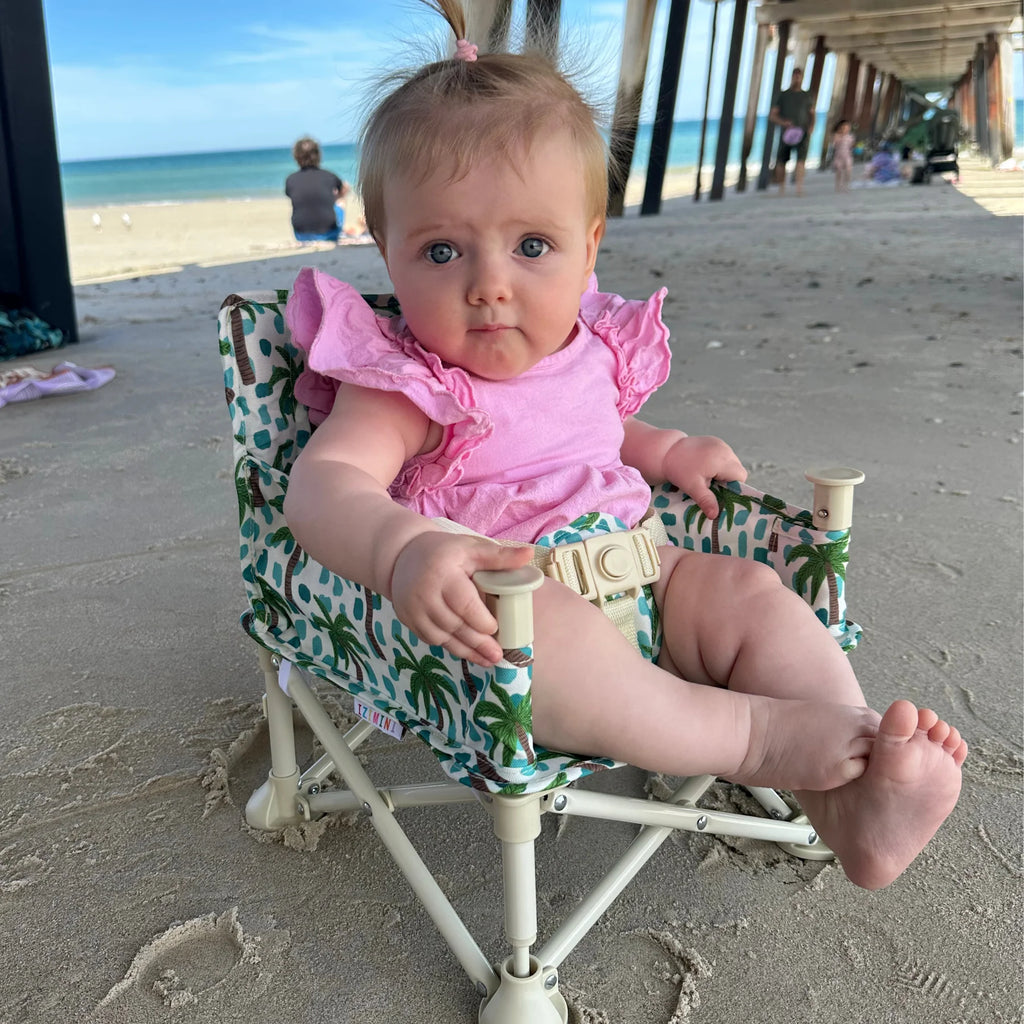 Baby sitting comfortably in Izimini Baby Chair Kai palm print chair at the beach – safe, portable and easy to clean.