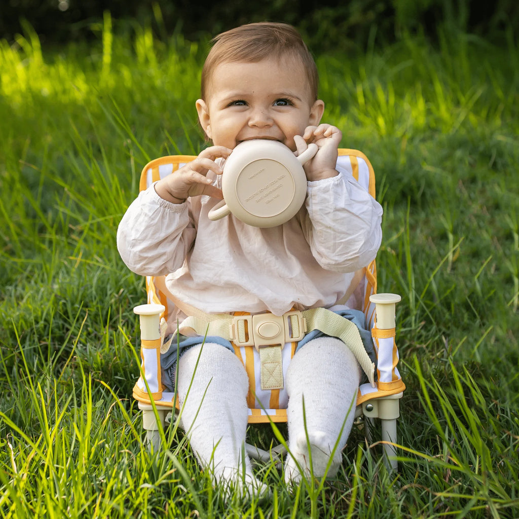 Baby sitting in the Izimini Brighton Baby Chair outdoors, holding a beige cup while seated on grass.