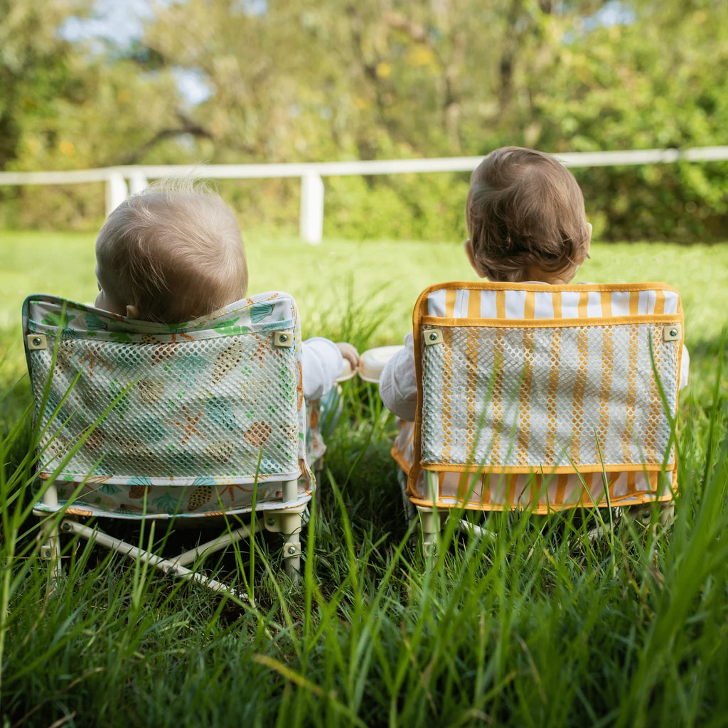 Two babies sitting outdoors in matching Izimini baby chairs, one in the yellow Brighton design, viewed from behind.