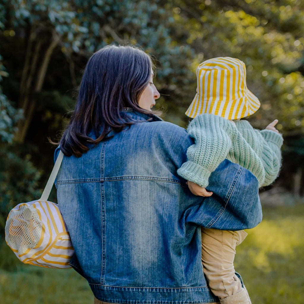 Parent carrying a baby wearing a yellow striped hat and matching Izimini Brighton baby chair bag outdoors.