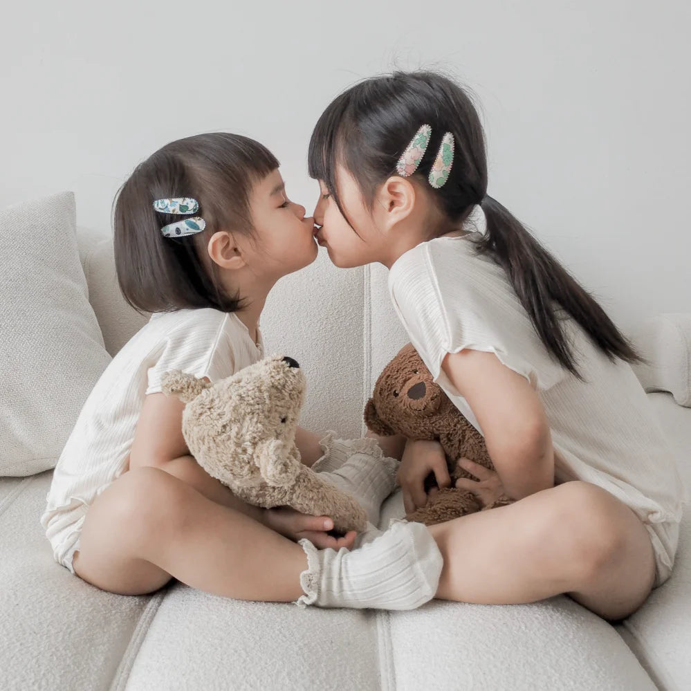 Two young children sitting on a couch, about to kiss, each holding a teddy bear.
