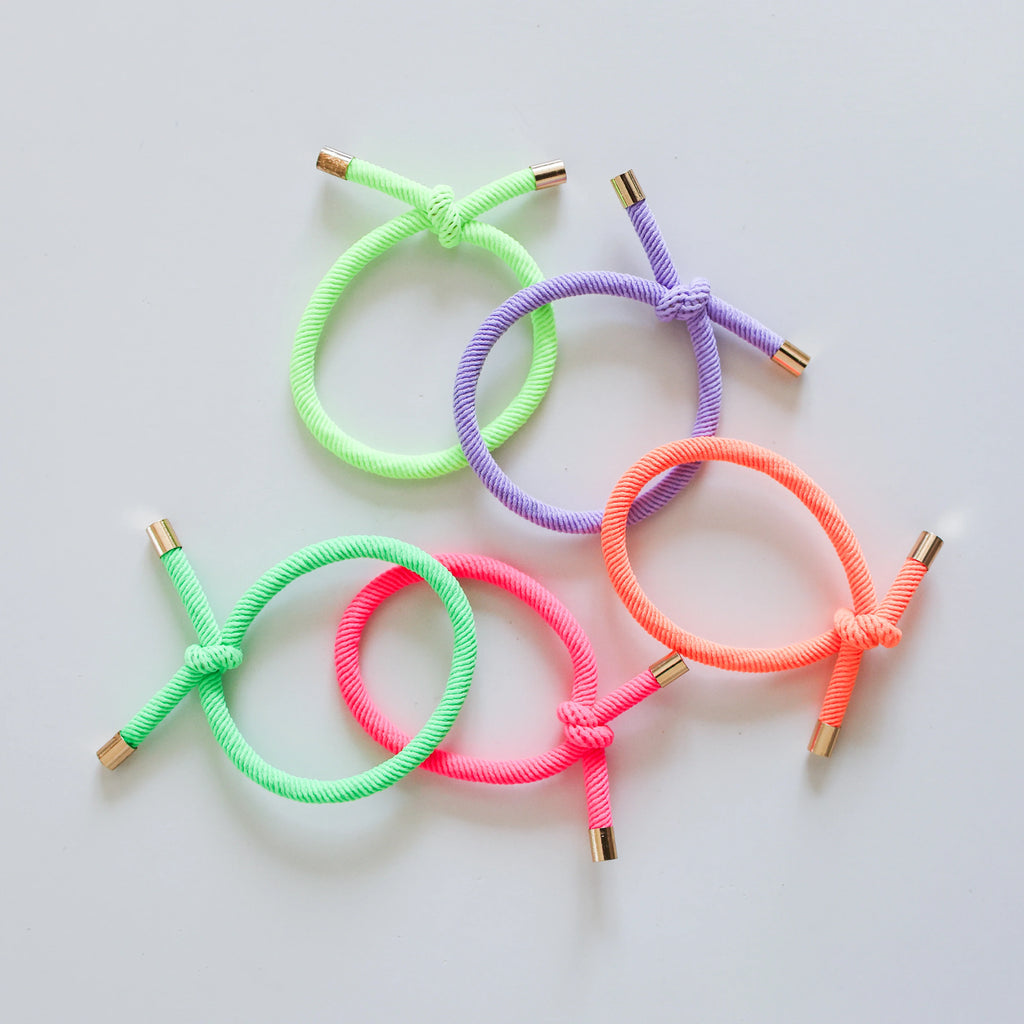 Five neon-coloured Josie Joan's hair ties laid flat, including green, purple, pink, and orange elastic ties with gold-tipped ends.