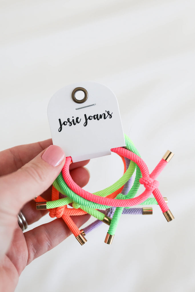 Hand holding a pack of Josie Joan's Neon Nora Hair Ties showing bright neon elastic bands and gold metal tips.