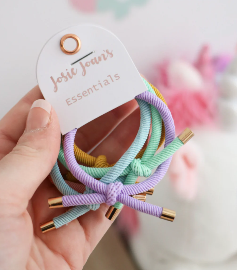 A set of five multicolored knotted hair ties with gold details, presented on a card with 'josie joans essentials' printed on it.