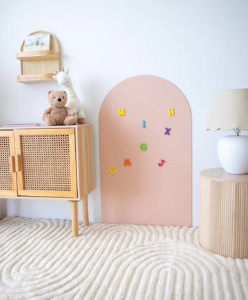 A blush MagPlay magnetic wall decal in an arched shape, styled in a child’s room with magnetic letters attached, positioned between a rattan cabinet and side table with a lamp.