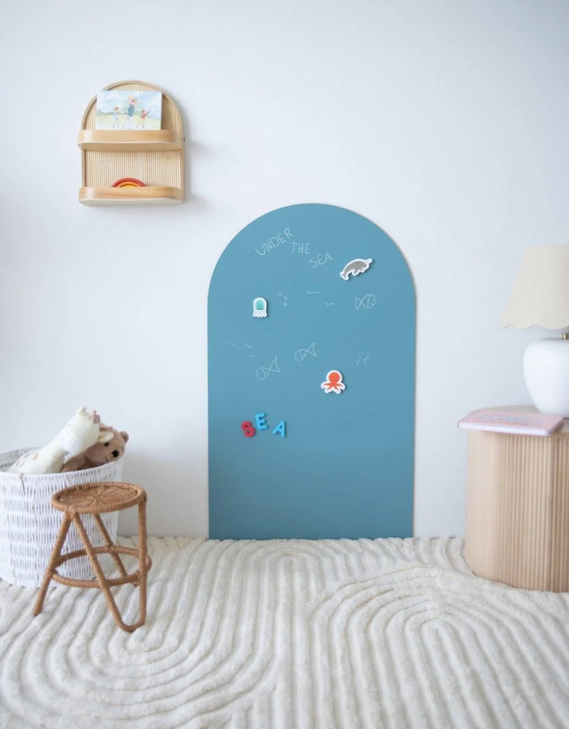 A teal MagPlay wall decal in an arch shape displayed in a kids' space, with magnets and chalk drawings applied, styled with soft furnishings and a rattan stool.