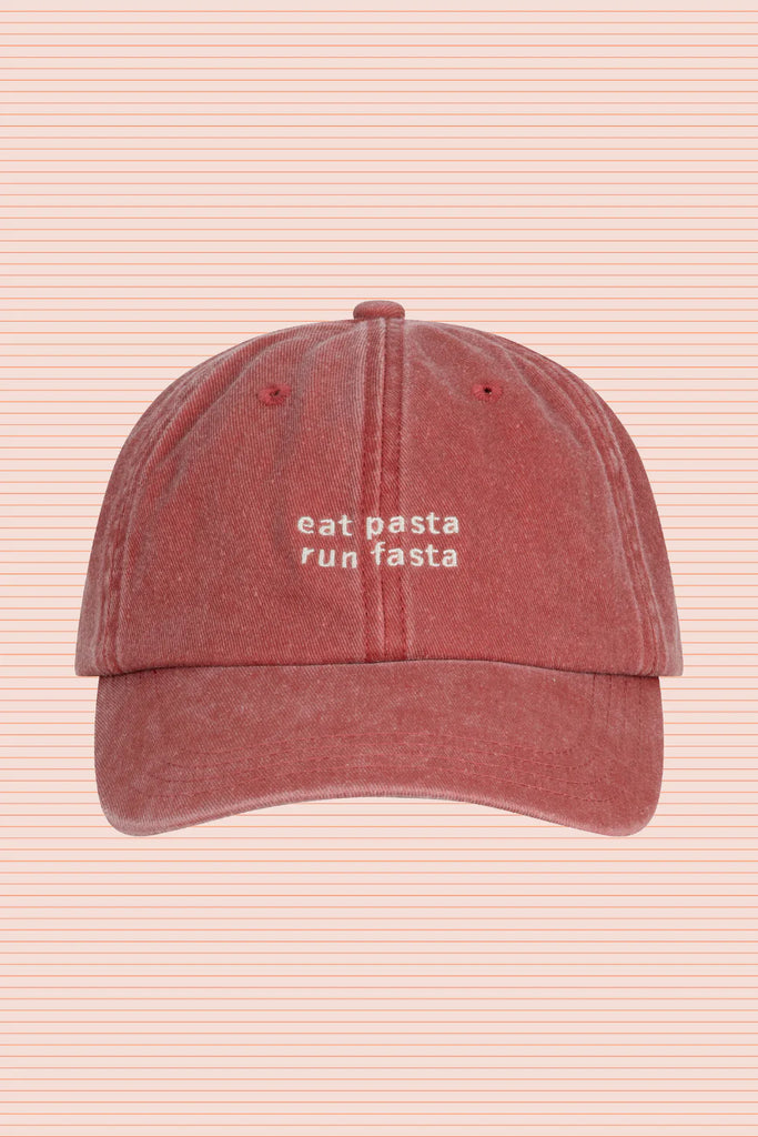 Soft cotton canvas cap in faded red with 'eat pasta run fasta' cream embroidery, displayed on a striped background.