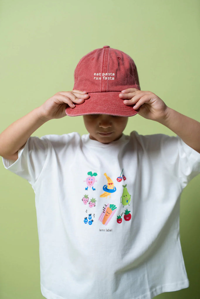 Child wearing faded red 'eat pasta run fasta' cap and white illustrated t-shirt, adjusting cap against a green backdrop.