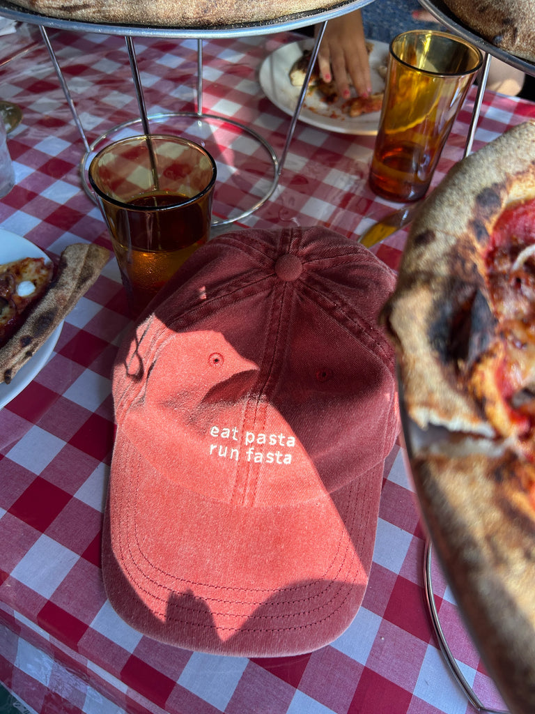Faded red 'eat pasta run fasta' cap placed on a checkered tablecloth surrounded by pizza and drinks.