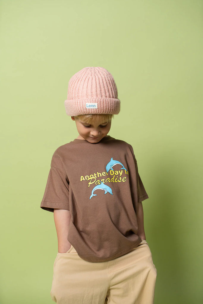 Young boy wearing the Paradise Tee in choc chip brown, paired with beige wide-leg trousers and a pale pink knit beanie, standing in front of a pale green backdrop. He smiles shyly, looking downward with hands in his pockets, highlighting the relaxed cut and soft cotton texture of the shirt.