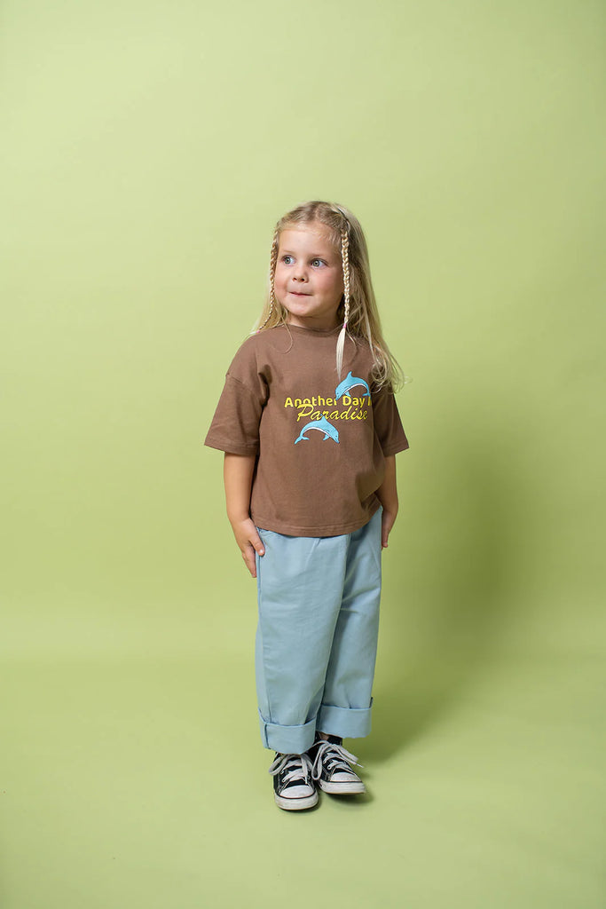 Young girl standing against a soft green backdrop, wearing the Paradise Tee in choc chip brown, styled oversized with light blue wide-leg pants and black Converse sneakers. She has long blonde hair and smiles slightly, looking off to the side, showcasing the loose fit and playful dolphin print.