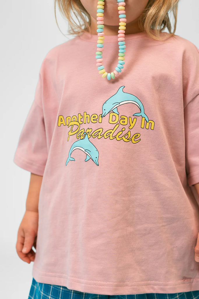 Close-up view of a child wearing the Paradise Tee in musk pink, showing the playful “Another Day in Paradise” text in yellow and script, with two light blue jumping dolphins across the chest. The child’s candy bead necklace and hands are visible, adding a playful and summery vibe.