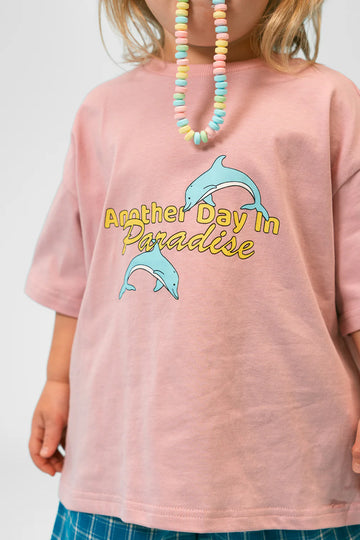 Close-up view of a child wearing the Paradise Tee in musk pink, showing the playful “Another Day in Paradise” text in yellow and script, with two light blue jumping dolphins across the chest. The child’s candy bead necklace and hands are visible, adding a playful and summery vibe.