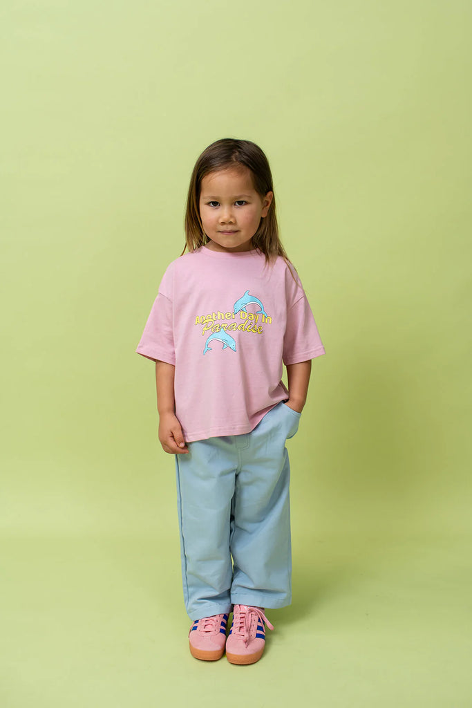 Young girl standing in front of a pale green backdrop wearing the Paradise Tee in musk pink, styled oversized with light blue pants and pink sneakers. She looks confidently at the camera, highlighting the tee’s loose, relaxed cut and dolphin print.