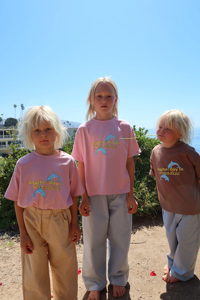 Three children standing outdoors on a sunny day, wearing the Paradise Tee in musk pink and brown. The two older girls wear the pink tee tucked into wide-leg pants, while the youngest child sports the brown version. They stand barefoot on a sandy path with greenery and a bright blue sky in the background, exuding a carefree, beachy vibe.