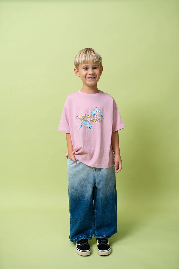 Young boy standing against a soft green backdrop wearing the oversized Paradise Tee in musk pink paired with wide-leg ombre blue jeans and black sneakers. He smiles casually, one hand in his pocket, showcasing the relaxed fit and playful front graphic.