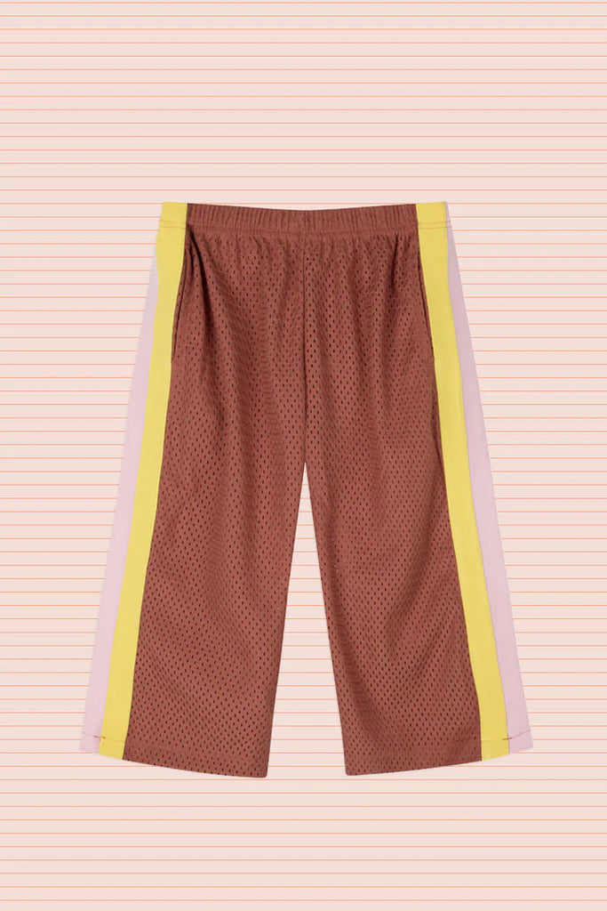 Flat lay of Sports Pant in Earth, displayed against a peach-striped background. The pants are made from rust-coloured perforated sports mesh with contrasting side stripes in soft pink and bright yellow, and feature an elastic waistband for easy fit.