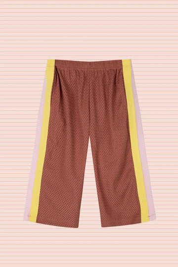 Flat lay of Sports Pant in Earth, displayed against a peach-striped background. The pants are made from rust-coloured perforated sports mesh with contrasting side stripes in soft pink and bright yellow, and feature an elastic waistband for easy fit.