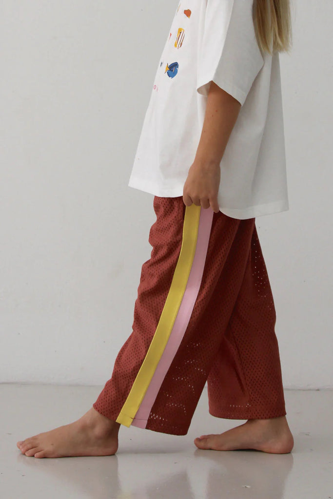 Side profile of child walking barefoot while wearing the Sports Pant – Earth, highlighting the full-length pink and yellow side stripes against the earthy rust mesh and the relaxed, comfortable fit.