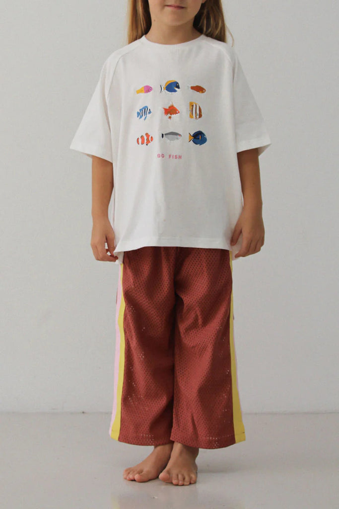 Child standing barefoot against a plain white wall, wearing the rust-coloured Sports Pant – Earth with yellow and pink side stripes, paired with a white oversized t-shirt featuring colourful fish illustrations.