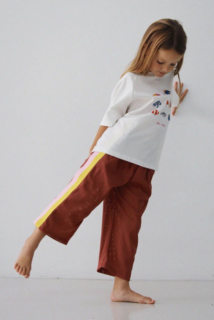 Child wearing the Sports Pant – Earth lifting one leg in a playful pose, showing the wide-leg cut and soft drape of the rust-coloured mesh fabric with bold yellow and pink side panels.