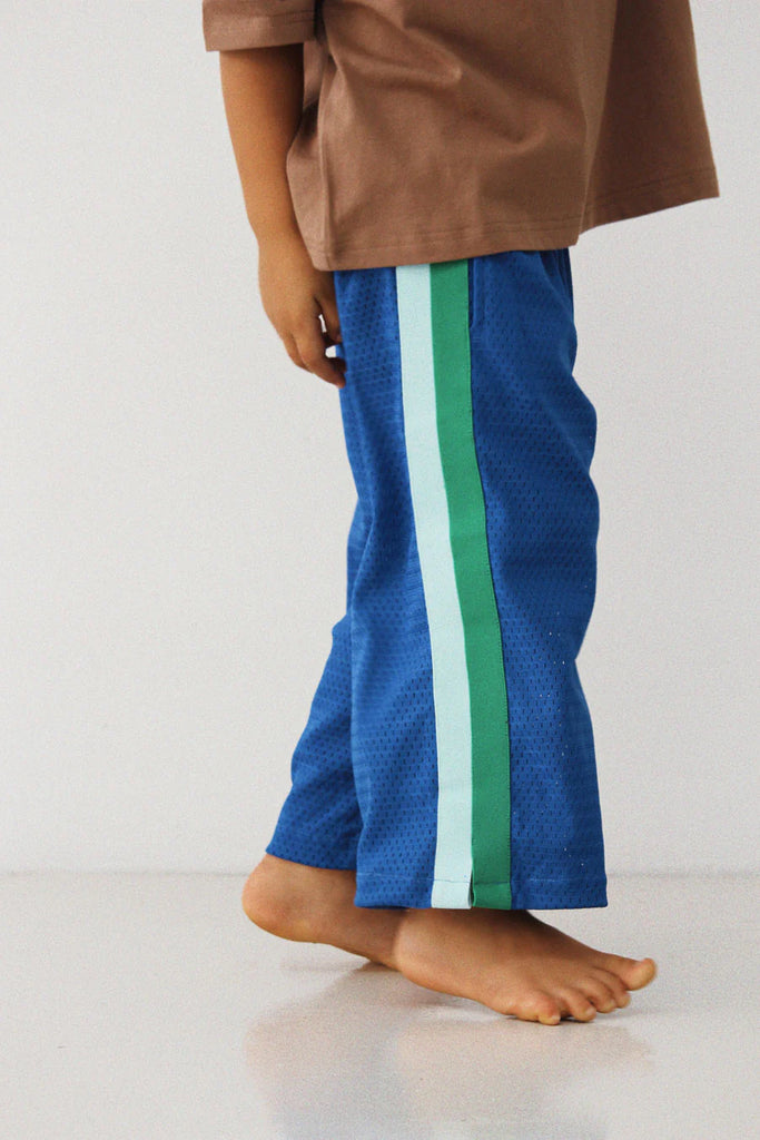 Close-up side profile of a child wearing the Sports Pant – Sea, showing the bright blue mesh fabric and the full-length seafoam green and pale blue side stripes. The child is barefoot, and the relaxed fit of the pants is visible.