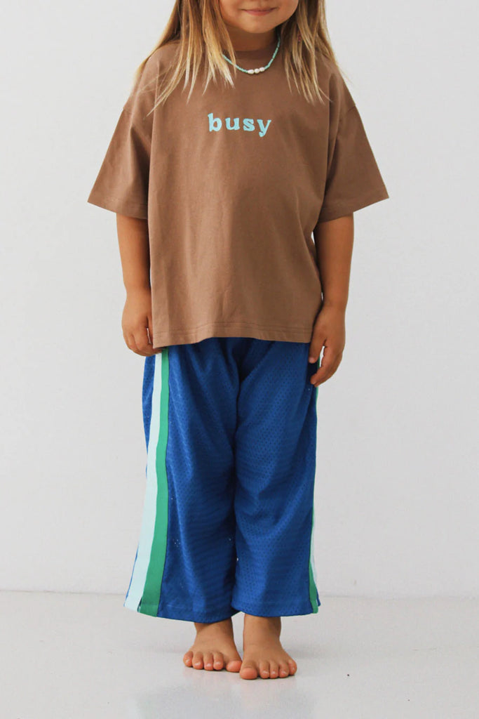 Side view of a child standing barefoot, wearing the Sports Pant – Sea with the brown “busy” tee, highlighting the relaxed cut and bold colour-blocked side stripe design.