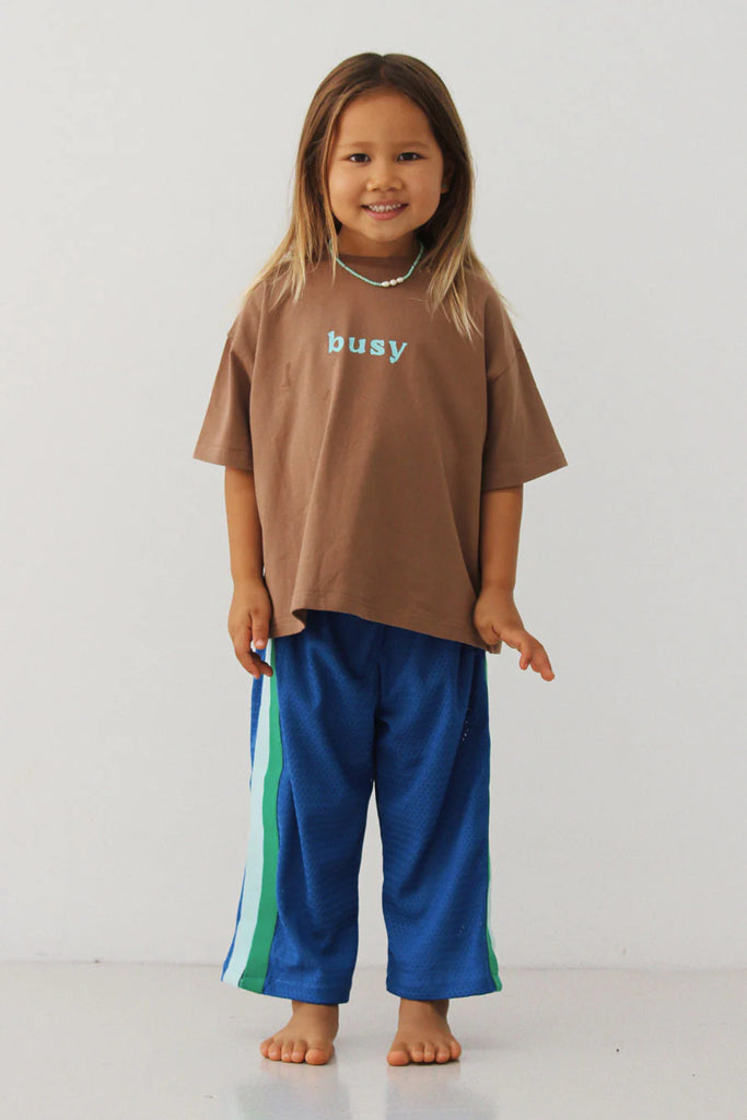 Smiling child standing barefoot against a plain white backdrop, wearing the cobalt blue Sports Pant – Sea paired with a brown oversized tee that reads “busy” in pale blue text, showing the playful colour contrast and wide-leg fit.