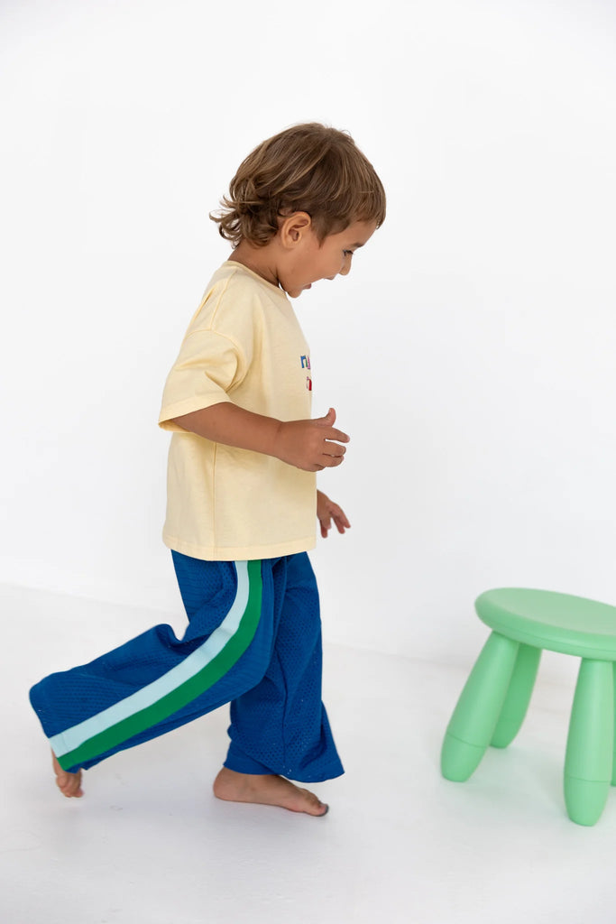 Child walking barefoot past a small green stool, wearing the cobalt blue Sports Pant – Sea paired with a pale yellow oversized t-shirt, capturing the motion-friendly, lightweight design and bright, playful colour palette.