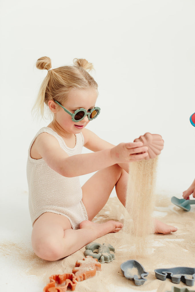 Sage green daisy-shaped kids’ sunglasses displayed on a neutral cream background.