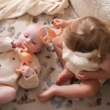 Two children playing with orange teething toys on a patterned rug.