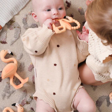Baby in a beige outfit with a teething toy, surrounded by toys on a patterned surface.