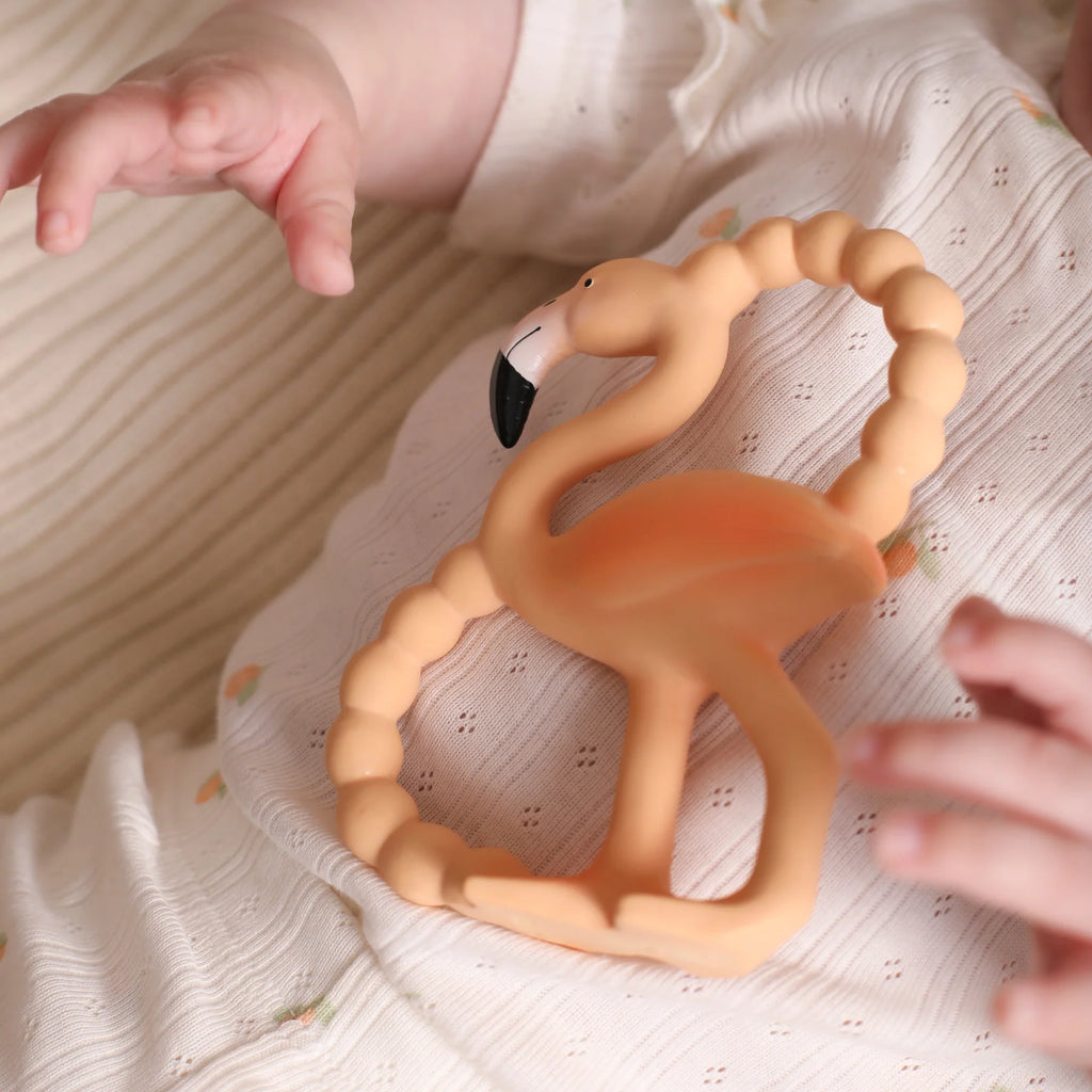 Flamingo-shaped teething ring on a textured surface with a baby's hand reaching towards it.