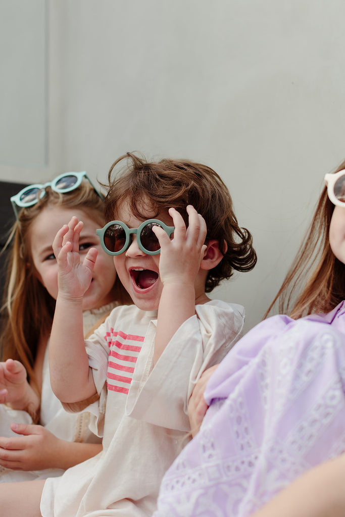 Sage green kids’ sunglasses with a rounded frame placed on a soft cream background.