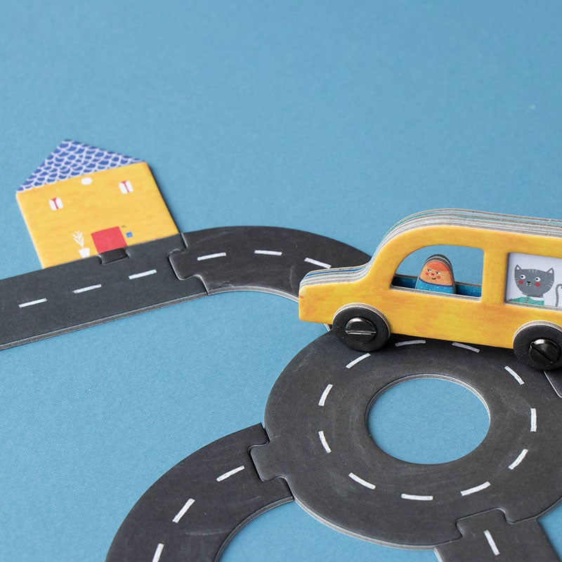 Yellow toy car driving on a connected road piece beside a small illustrated house from the Londji Roads Pocket Game.