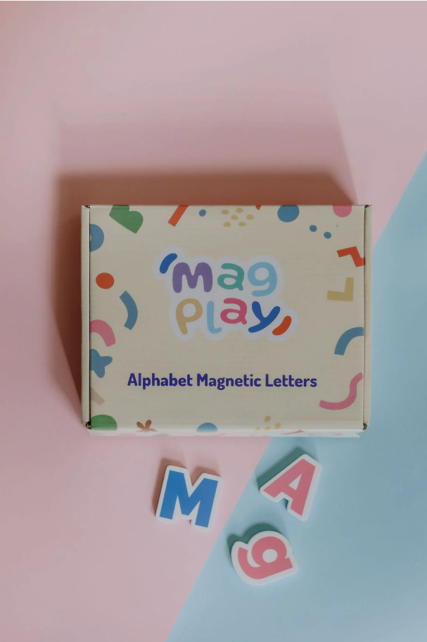 Flat lay of the MagPlay Alphabet Magnetic Letters box on a pink and blue background, with magnetic letters spelling “MAG” scattered nearby.