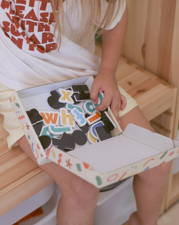 Close-up of a child sitting and playing with a box of colourful MagPlay alphabet magnets on their lap.
