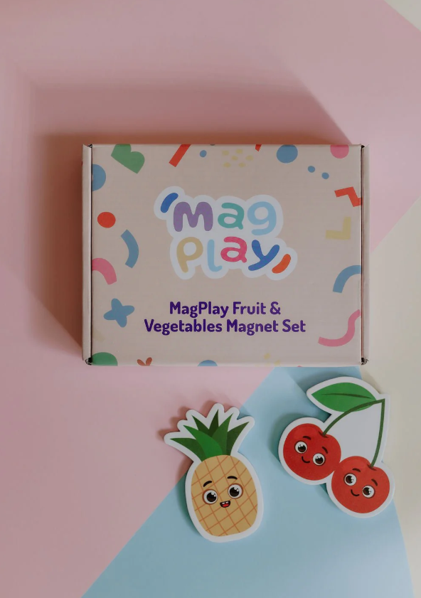 Top-down view of the MagPlay Fruit & Vegetables Magnet Set box on a pastel pink and blue background with pineapple and cherry magnets beside it.