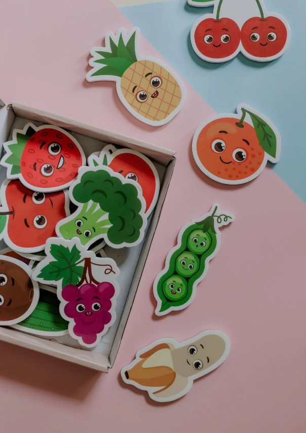 Flat lay of the open MagPlay box showing colourful fruit and vegetable magnets including peas, grapes, and tomato, styled on a pink background.