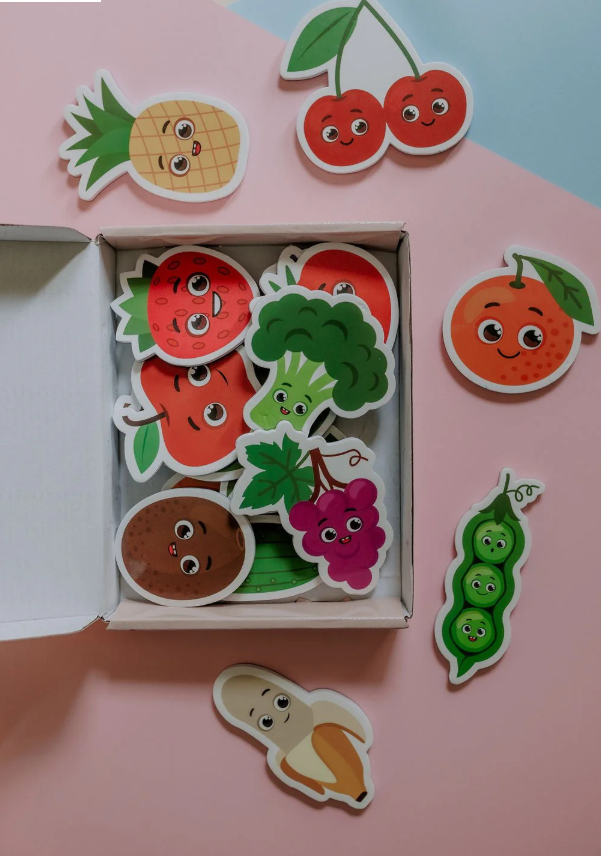 Top-down view of the MagPlay magnet kit with pieces like a banana, broccoli, and cherry displayed around an open box on a pastel backdrop.