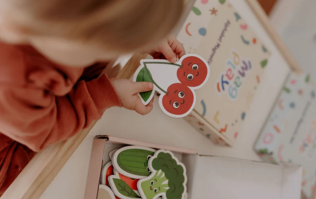 Child reaching into the MagPlay Fruit & Vegetables Magnet Kit box, selecting a cherry magnet during playtime at a table.