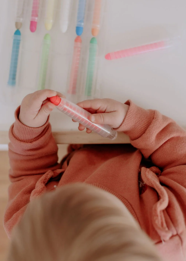 Toddler in pink holding a red crayon from the set while seated at a table.
