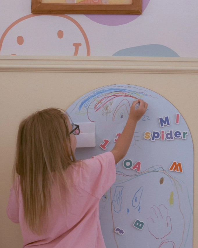 Young child using the crayons to draw on a vertical surface with magnetic letters in the background.