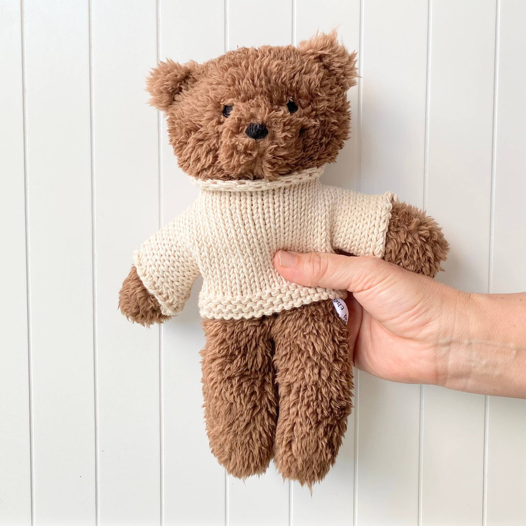 Hand holding Milo Bear Mini soft toy made from chocolate brown plush fabric with cream knitted merino wool jumper against white panel wall.