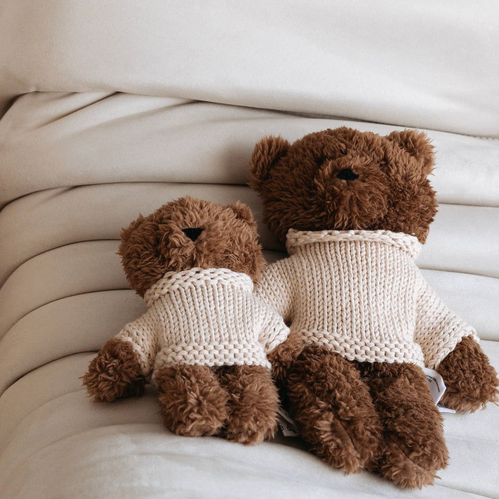 Milo Bear Mini soft toy lying on a cream bedspread next to medium-sized Milo Bear, both wearing cream knitted merino wool jumpers.