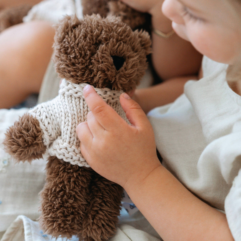 Child cuddling Milo Bear Mini soft toy made from chocolate plush fabric, dressed in a cream merino wool jumper, sitting on a white bed.
