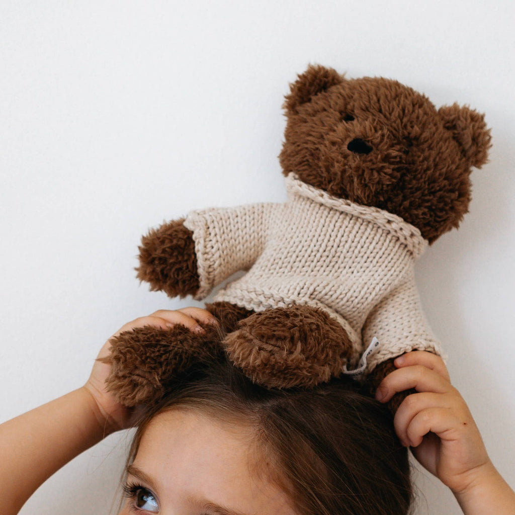 Child holding Milo Bear soft toy on top of their head, showing chocolate brown plush fur and cream knitted merino wool jumper.